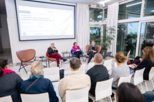 Ujęcie sceny, na której zasiada troje panelistów, dwie kobiety oraz jeden mężczyzna. Siedzą na dużych skórzanych fotelach, obok nich jeden fotel jest pusty. Za nimi rzutnik z prezentacją. Widać rózniez zgromadzona publiczność od tyłu.