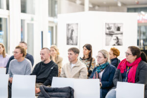 Publiczność podczas seminarium. Osoby w różnym wieku, wśród nich kobiety i mężczyźni, wpatrują się w stronę sceny. Sala w której się znajdują jest jasna, przestronna, w wieloma przeszkleniami. W tyle widać fragmenty wystawy fotograficznej.