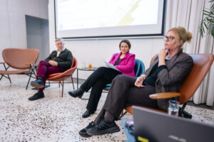 Troje panelistów, w tym dwie kobiety i jeden mężczyzna siedzą przed publicznością zgromadzona na seminarium. Za nimi na rzutniku wyświetlana jest prezentacja.