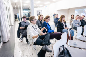 Zbliżenie na publiczność podczas seminarium. Na pierwszym planie kobieta w krótkich jasnych włosach mówi, gestykulując. Jest zwrócona w stronę panelistów, znajdujących się poza kadrem.
